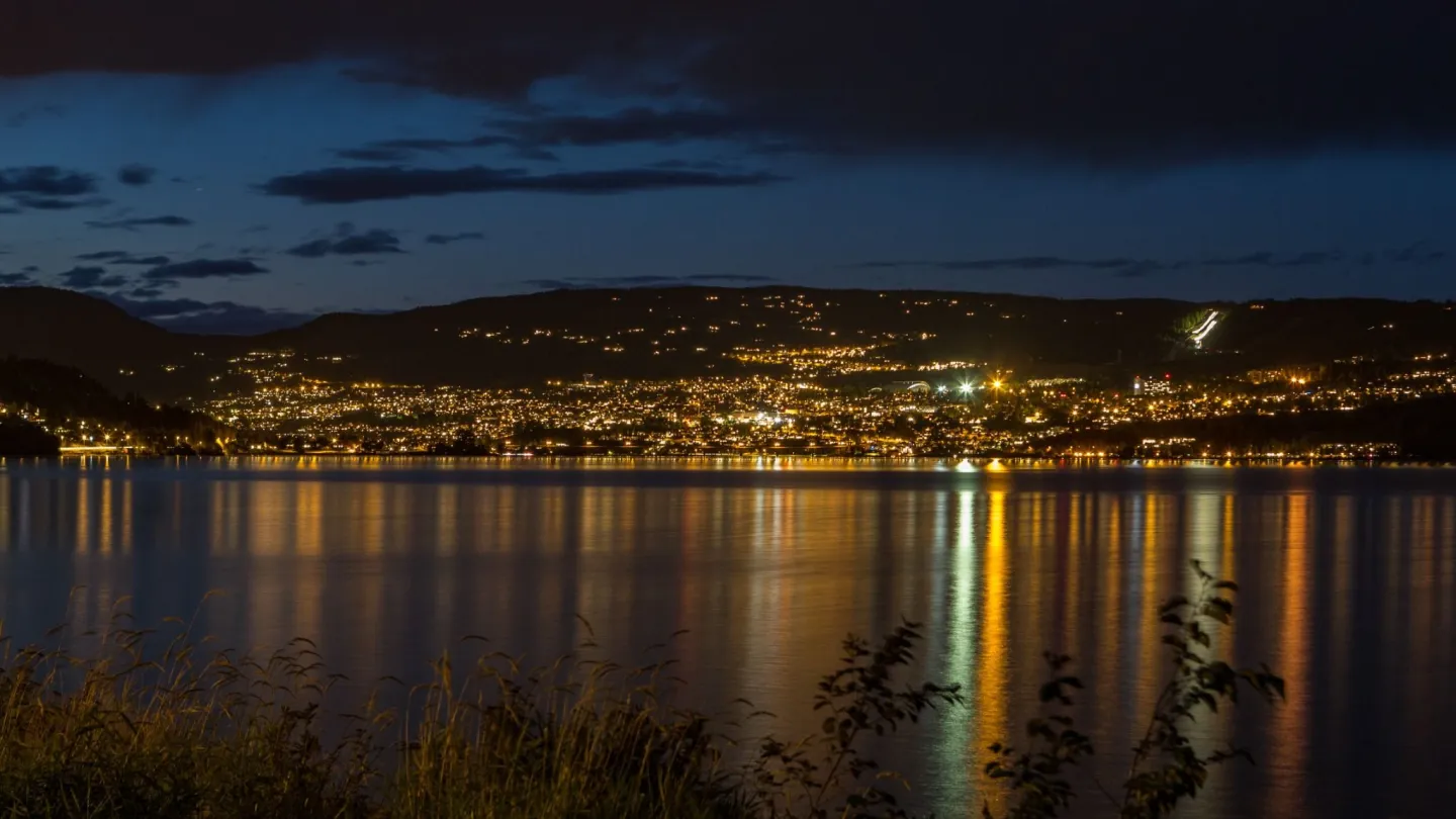 Kveldsutsikt over Lillehammer med byens lys reflektert i Mjøsa, sett fra bredden, Perfekt stemningsbilde for sommerferieopplevelser og avslappende overnatting